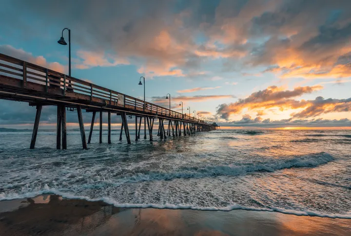 The Imperial Beach Pier reaching out over the surf at sunset, its lamps lit and the sky orange behind it.
