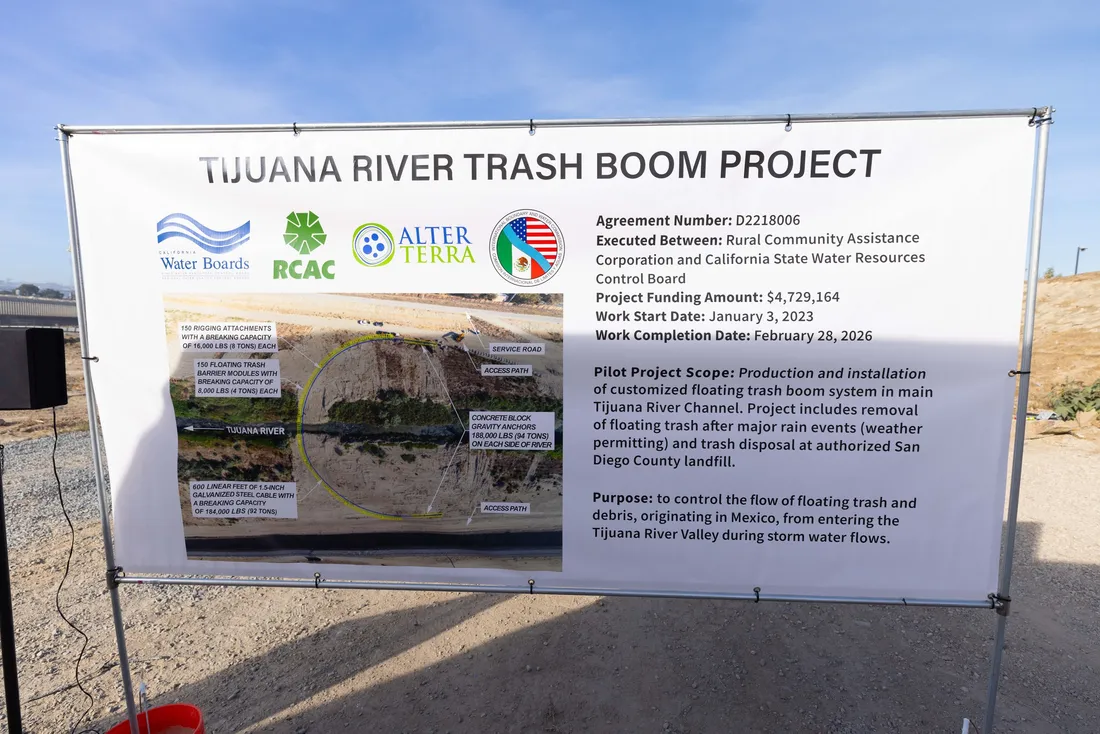 A project signboard on open ground reading TIJUANA RIVER TRASH BOOM PROJECT, carrying the Water Boards, RCAC and Alter Terra logos, an aerial diagram of the boom stretched across the river channel, and a panel of project details.